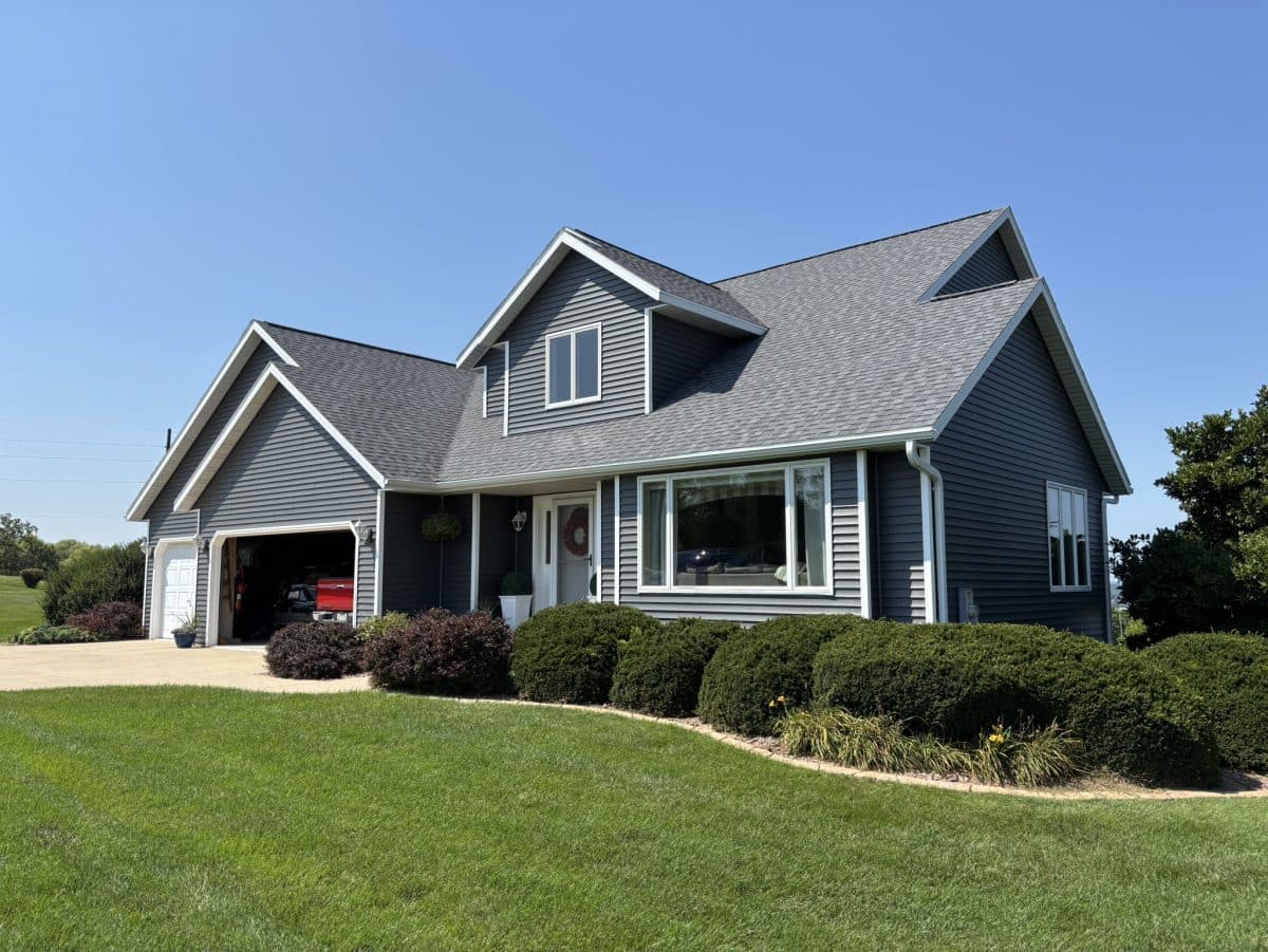 A modern gray house with white trim and roofing Deforest expertise, featuring a two-car garage, well-maintained bushes and lawn under a clear blue sky. The garage door is open, revealing a red vehicle inside.