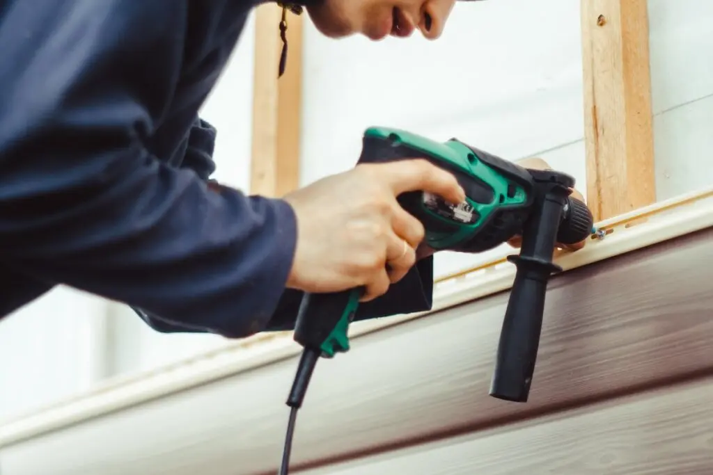 siding installation A person using a green and black power drill to fasten a screw into a wooden frame, working on construction or renovation near an exterior wall.
