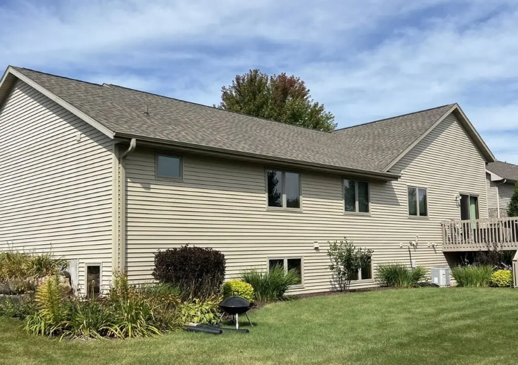 A beige, two-story suburban house with a sloped roof, small windows, and a wooden deck, surrounded by green grass, shrubs, and plants under a partly cloudy sky.