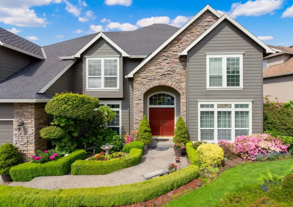 siding installation A two-story house with gray siding and stone accents, large windows, a red double front door, and a well-kept garden with trimmed shrubs, flowers, and a curved walkway under a blue sky with scattered clouds.