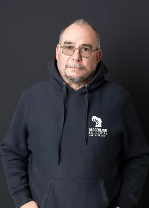 A middle-aged man with glasses and a beard stands against a dark background, wearing a navy blue hoodie with a 