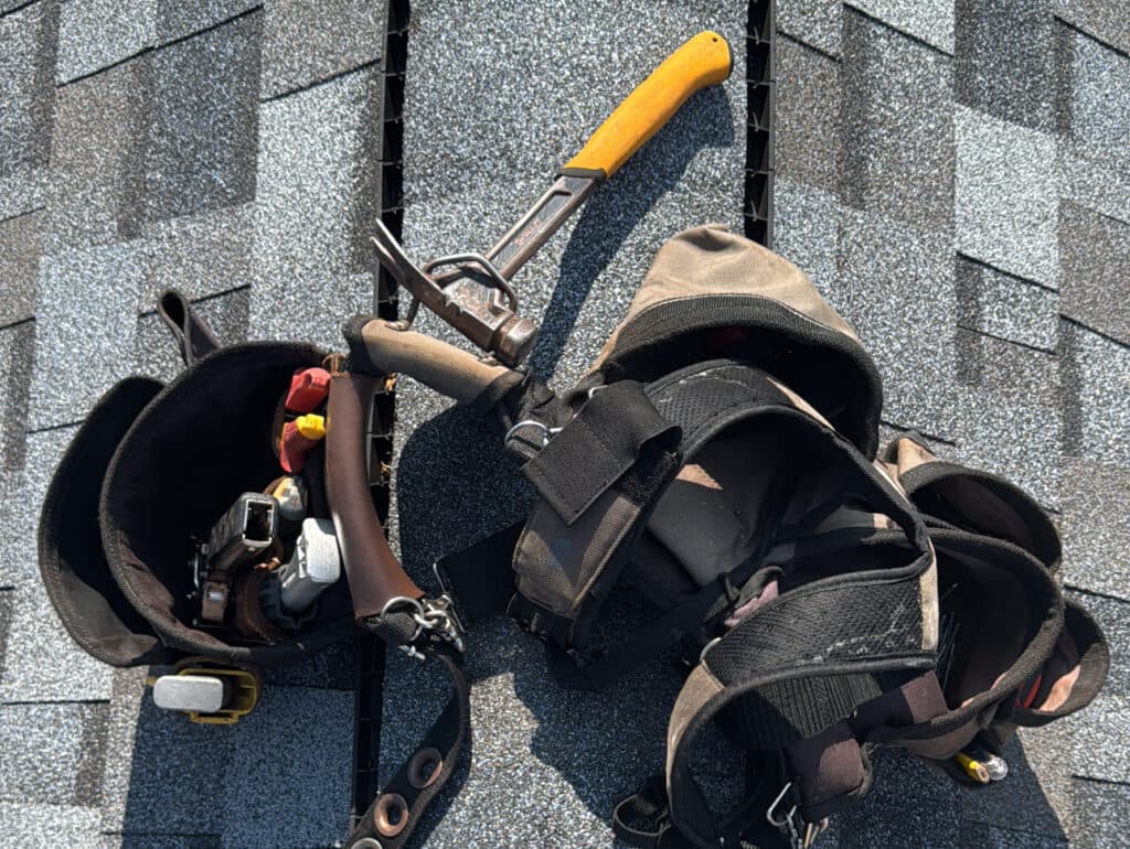 roof maintenance checklist Two tool belts with various roofing tools and a yellow-handled snip sit on gray shingle rooftop under bright sunlight.