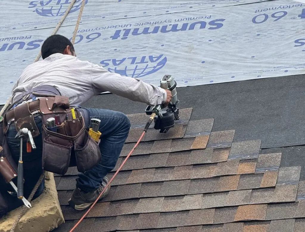 A construction worker wearing a tool belt installs asphalt shingles on a roof using a nail gun. Roofing underlayment and materials are visible in the background.