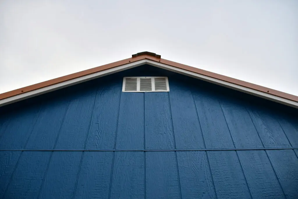 The image shows the upper portion of a blue building with a triangular roof, brown trim, and a small white vent near the roof peak against a pale sky.