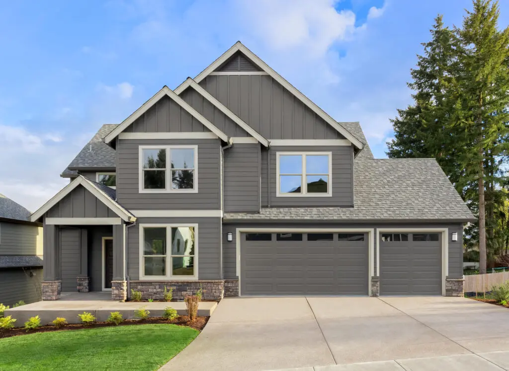 A modern, two-story gray house with a gabled roof, large windows, and a three-car garage. The front yard features a neatly manicured lawn and small shrubs. The sky is partly cloudy, and tall trees are visible in the background.