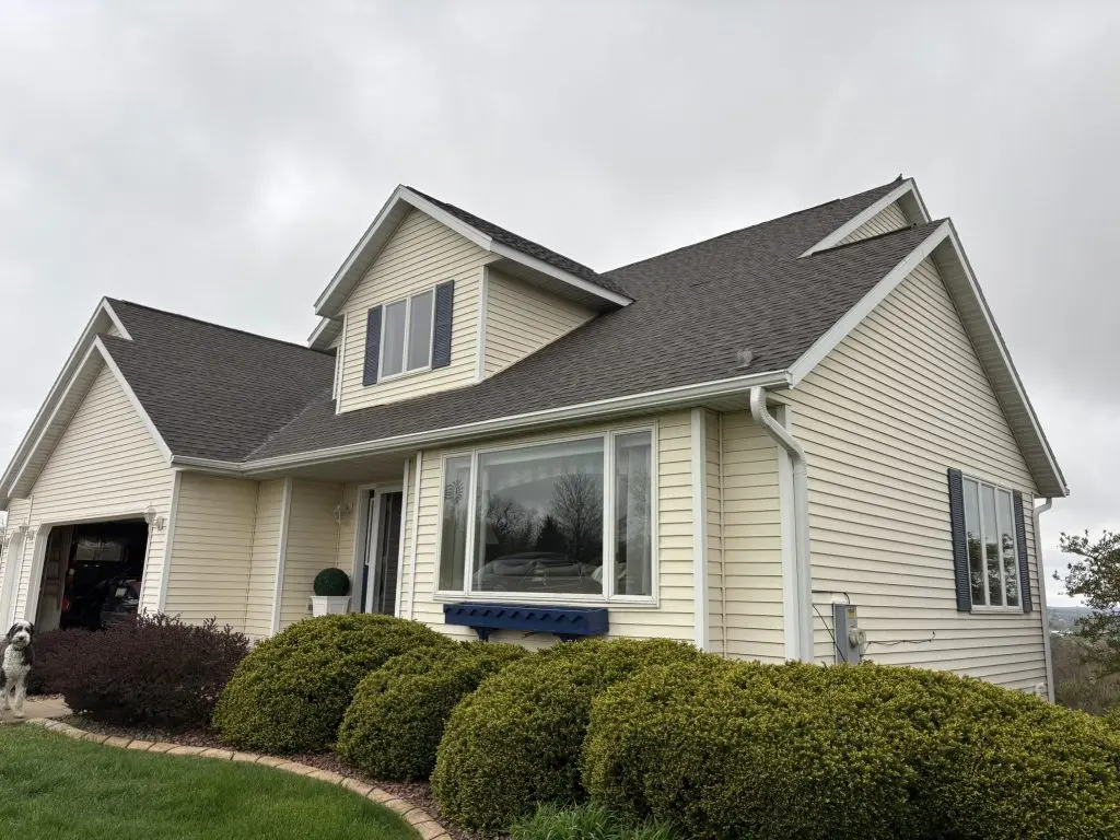 A beige, two-story suburban house with dark shingles, white trim, large windows, bushes in front, and a cloudy sky in the background.