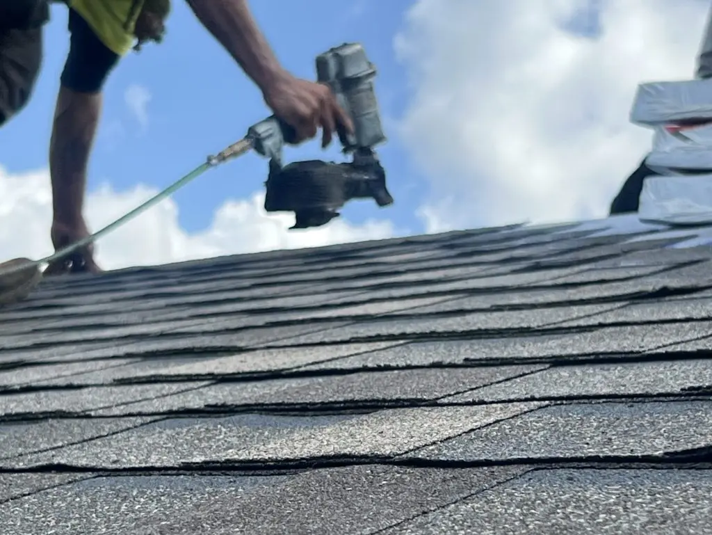A worker in a yellow vest uses a nail gun to install asphalt shingles on a roof under a partly cloudy sky.