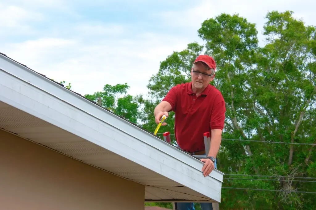 A man wearing a red shirt and cap stands on a ladder, cleaning leaves from a house gutter with a yellow tool. Green trees and a cloudy sky are in the background.