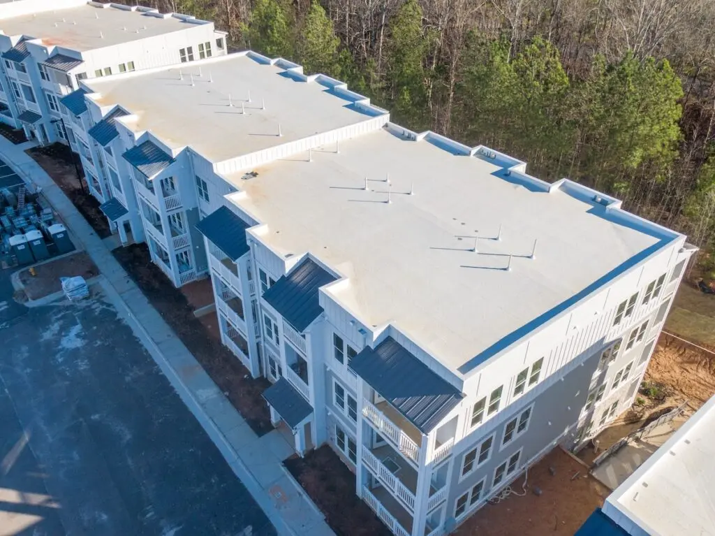 Aerial view of a modern, three-story apartment building with a white commercial roof type and exterior, surrounded by trees and an unfinished area scattered with dirt and construction materials. commercial roof types
