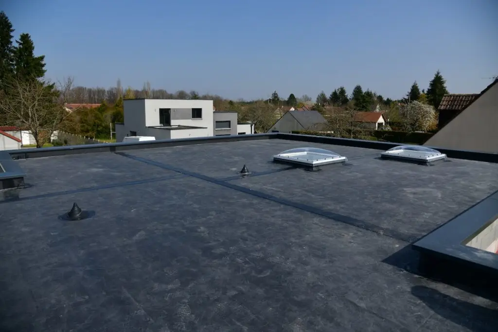 Flat black rubber roofing with two square skylights and several vents, overlooking a suburban neighborhood filled with modern and traditional houses, trees, and a clear blue sky.