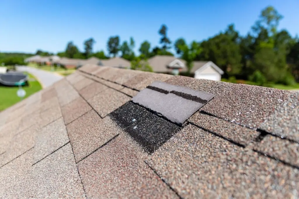Close-up of a house roof with missing shingles on roof, highlighting one damaged or absent shingle that exposes the black underlayment. Neighboring houses and trees appear blurred in the background.