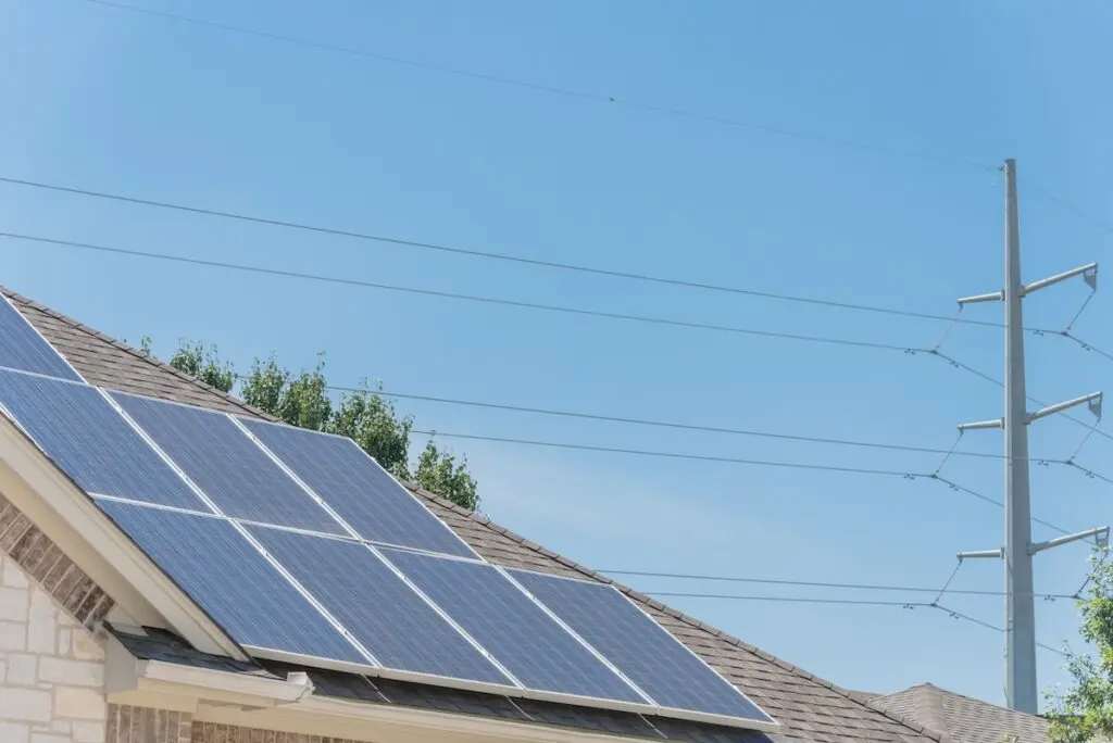A rooftop with solar panels under a clear blue sky. Nearby, an electrical pole with power lines is visible. Trees are partially seen in the background, indicating a suburban setting.