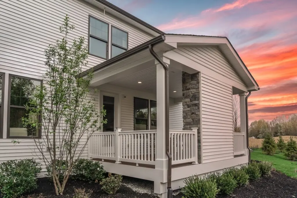 A modern house with beige siding and a small covered front porch with white railings, set against a colorful sunset sky. There are shrubs and a tree planted along the landscaped front yard.
