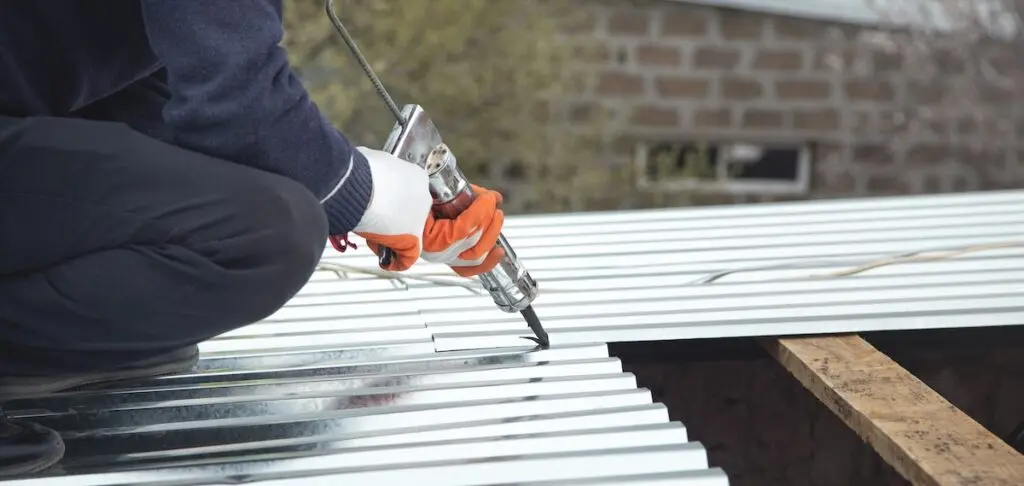 waterproof sealant for metal roof A person wearing orange gloves and using an electric drill is installing or securing metal roofing sheets on a building, likely applying waterproof sealant for metal roof protection. Part of a brick wall and some greenery are visible in the background.