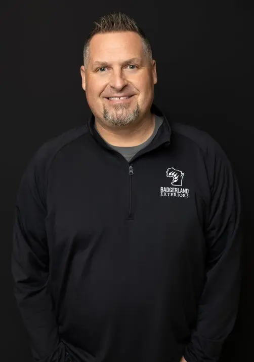 A man with short hair and a trimmed beard smiles at the camera, wearing a black zip-up shirt with the 