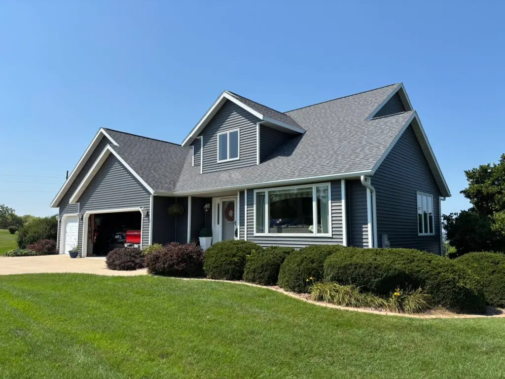 A modern gray house with white trim and roofing Deforest expertise, featuring a two-car garage, well-maintained bushes and lawn under a clear blue sky. The garage door is open, revealing a red vehicle inside.