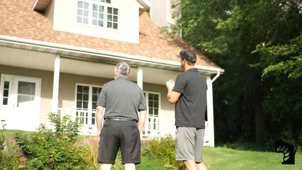 Two men stand in a grassy yard, gazing at a beige house with a sloped roof and white windows. One man is older with gray hair, the other younger, wearing a cap. Trees frame the scene on the right, perfectly setting up for a solar installation in Waunakee.