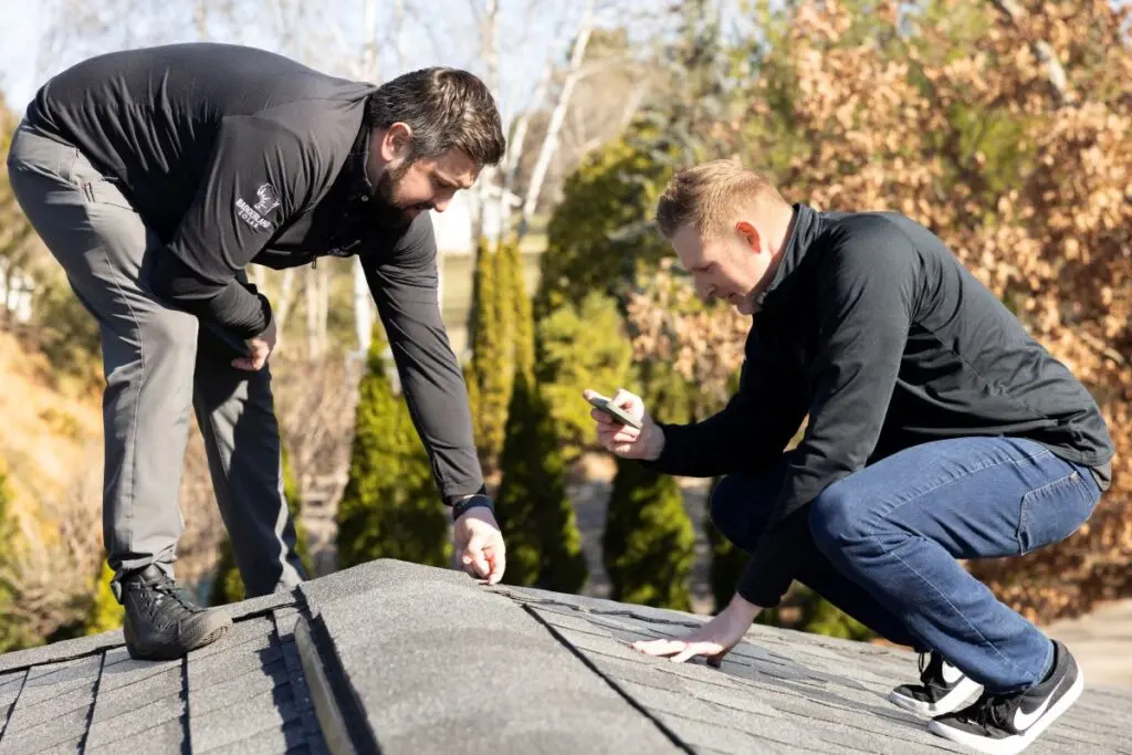 wind damage roof repair Two men on a rooftop inspect shingles; one crouches while taking a photo with a smartphone, and the other bends down to closely examine the roof surface. Trees and blurred nature are visible in the background.