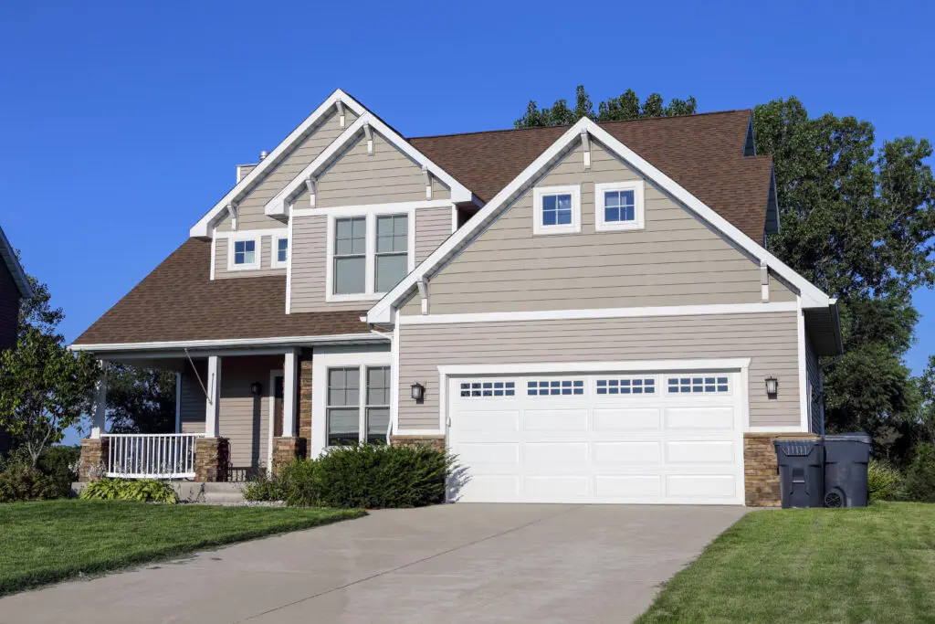 siding insulation A modern two-story house with taupe siding, white trim, and a brown roof, featuring a large white garage door and front porch. Enhanced siding insulation ensures energy efficiency, while a well-kept lawn shines under a clear blue sky.