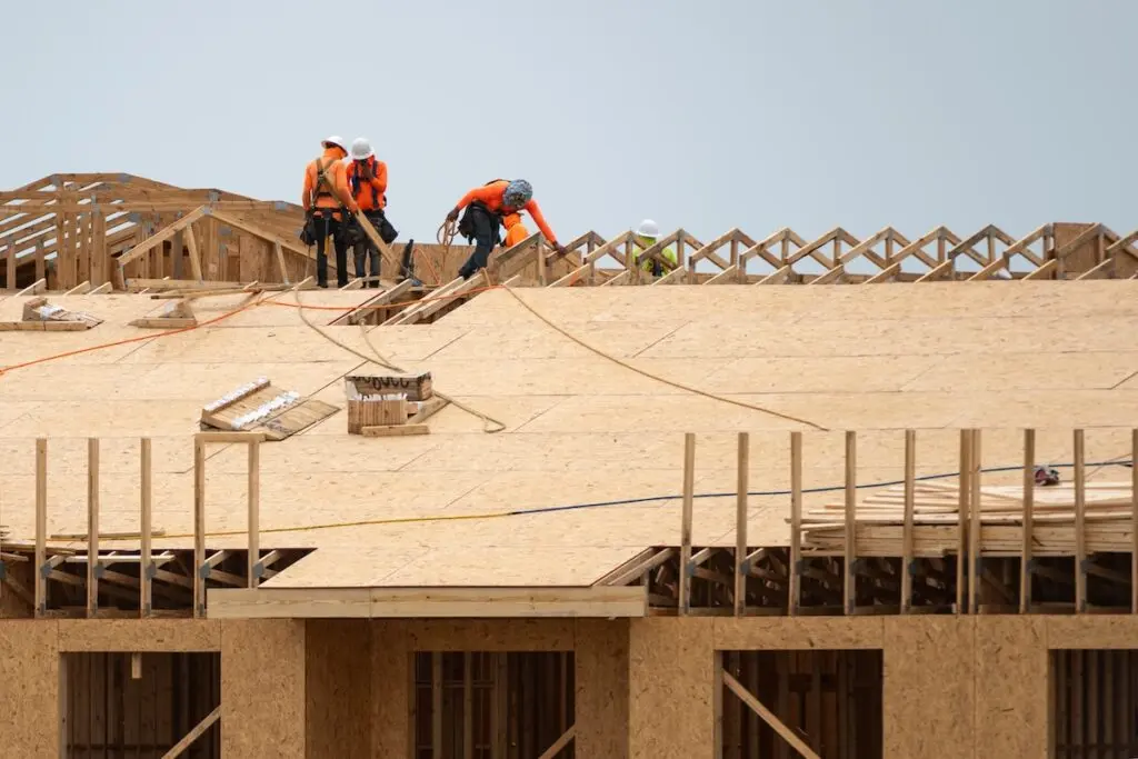 Construction workers wearing safety gear are assembling the wooden framework of a roof on a building. They are dressed in orange and are positioned on top of the structure, surrounded by wooden beams and panels under a clear sky.