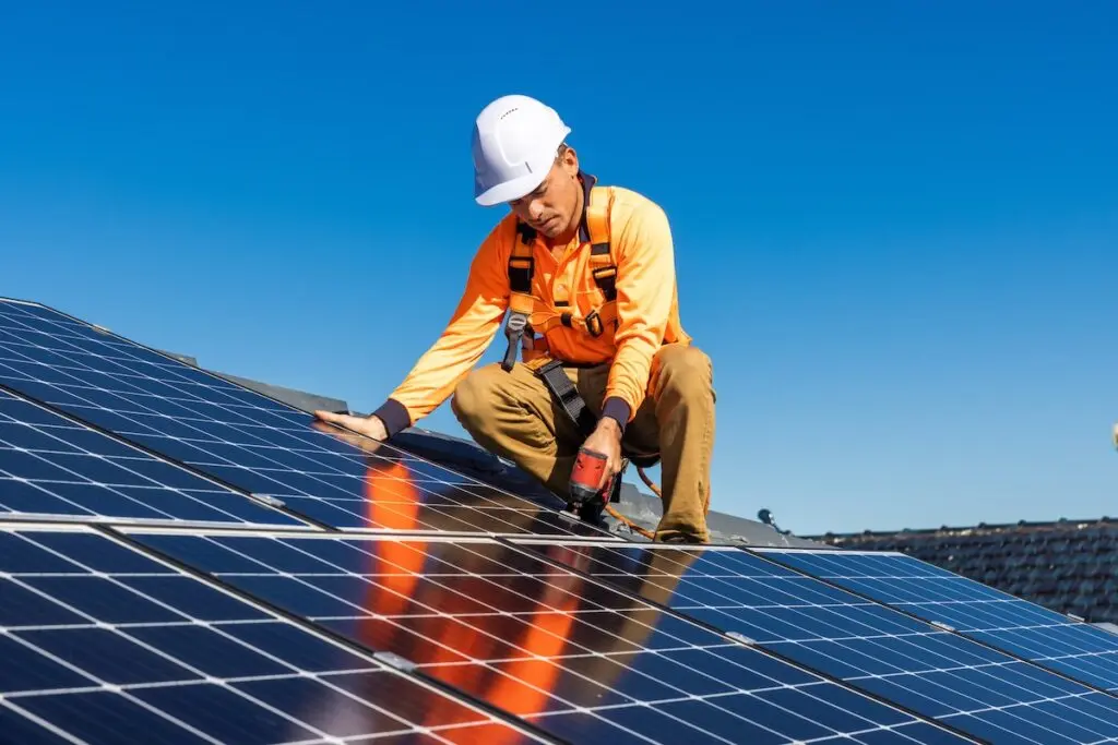 A worker in an orange shirt and white hard hat is installing solar panels on a roof. He is crouched down, using a tool on the panels, against a clear blue sky backdrop. Safety harnesses and ropes are visible.