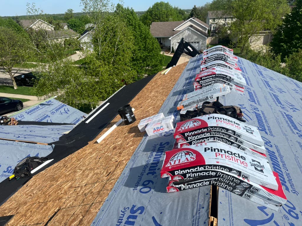 roofing companies sun prairie wi Roof under construction with stacks of shingles, tools, and materials placed on a partially completed roof. Surrounding trees and houses are visible in the background under a clear sky.
