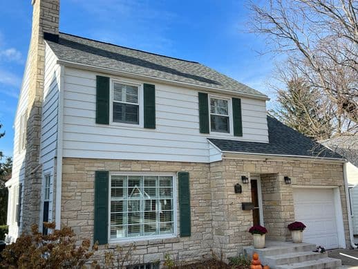 A two-story house with stone and white siding, green shutters, and a chimney. The front steps are decorated with potted plants and a small pumpkin. The sky is clear and blue.