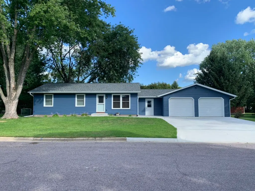 A single-story blue house with white trim features a new roofing Oconomowoc style, a large lawn, and a driveway to a double garage. Trees surround the property under a partly cloudy sky.