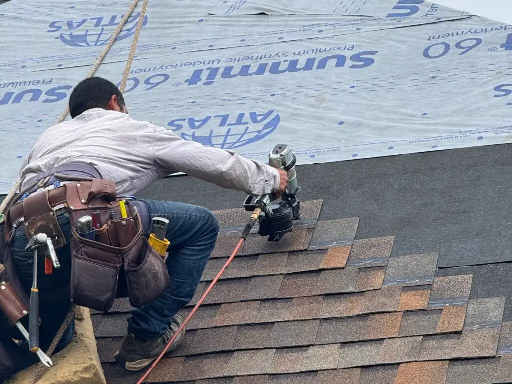 asphalt shingle roof replacement A construction worker in a tool belt uses a nail gun to install asphalt shingles on a roof, while another worker in red sits higher up, securing roofing underlayment.