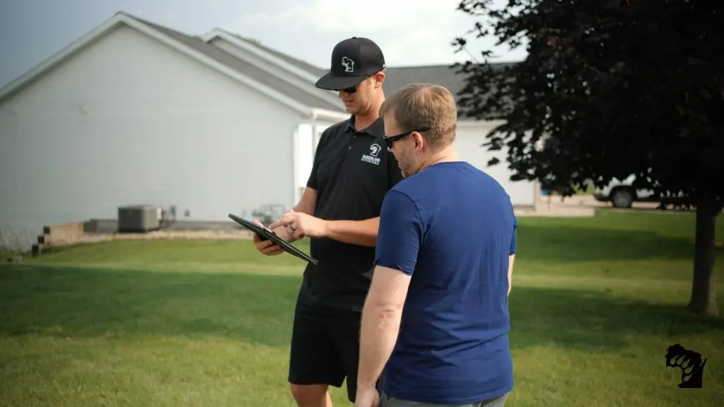 Two men stand on a lawn; one in a black outfit shows something on a tablet to the other in a blue shirt. A white house and tree are in the background.