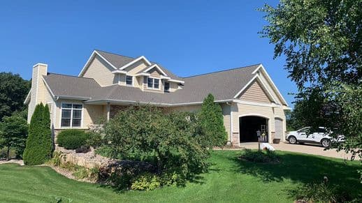 A beige two-story house with a gray roof and white trim sits under a clear blue sky. The front yard is landscaped with shrubs and a lush green lawn. A driveway leads to an open garage, where a white car is parked.