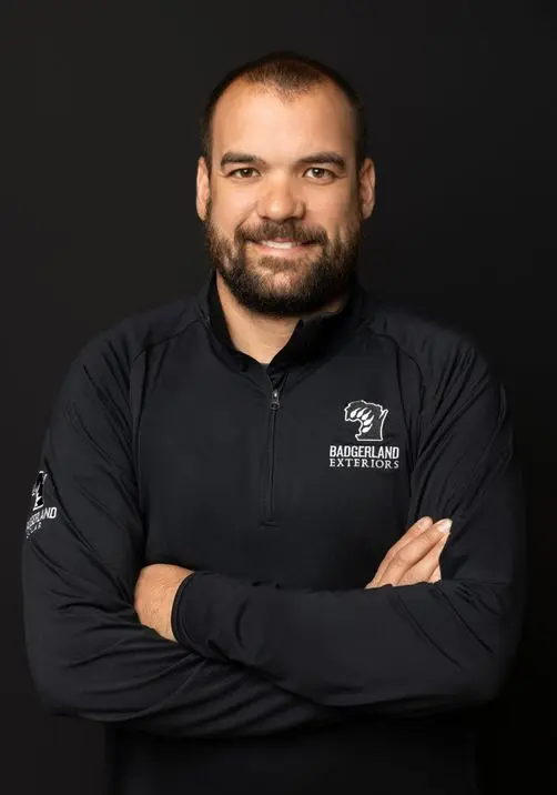 A man with a beard and short hair stands against a dark background, smiling with his arms crossed. He is wearing a black zip-up shirt with the 