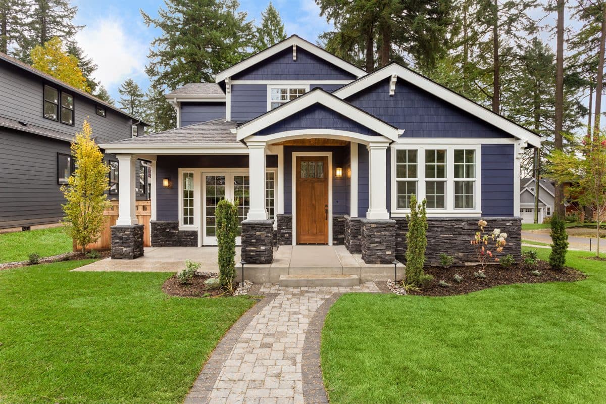 A charming modern house with blue siding, white trim, and a stone walkway. It features a wooden front door, multiple windows, and a well-kept lawn surrounded by tall trees on a sunny day.