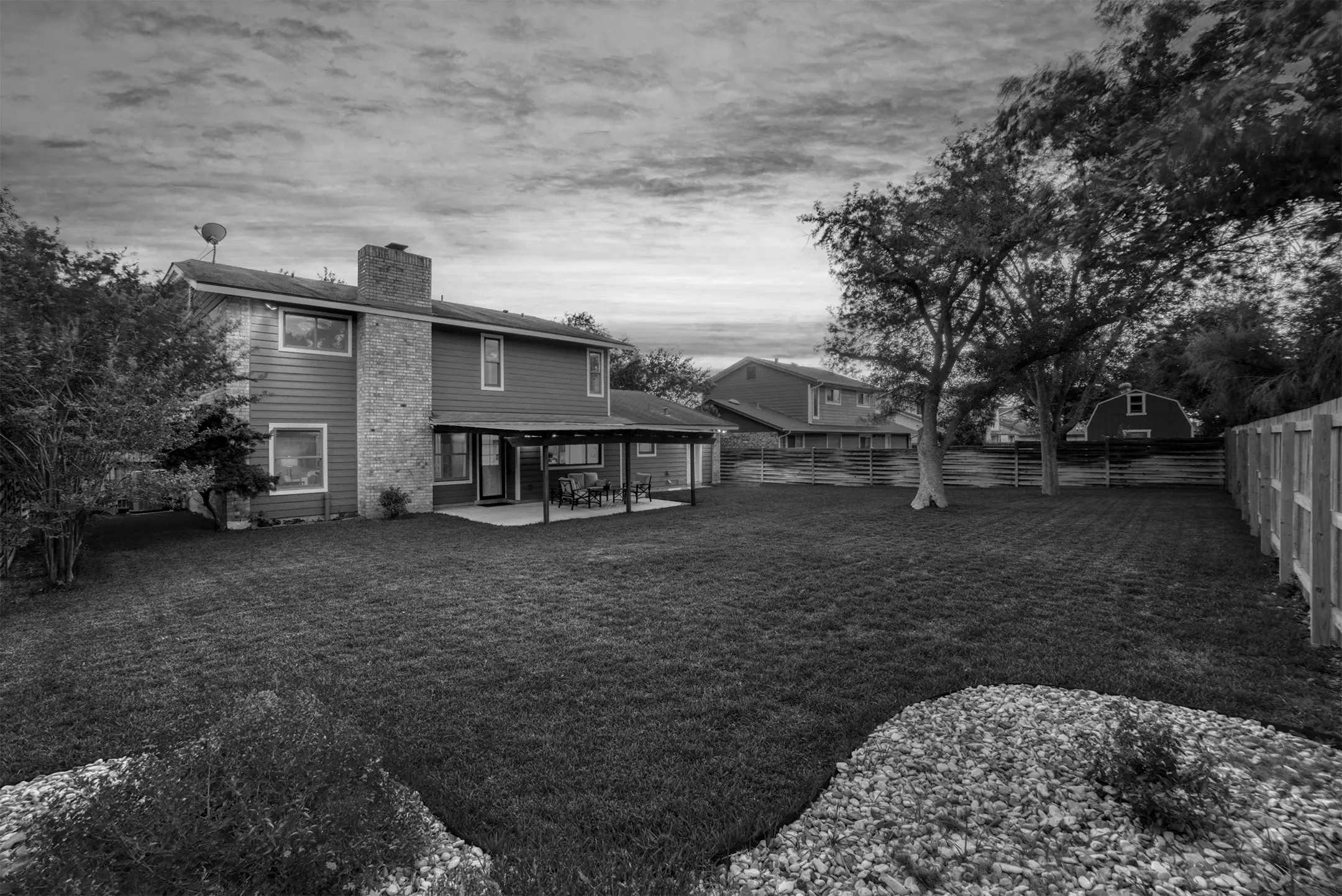 Black and white photo of a suburban backyard with a two-story house. The yard features a spacious lawn, a covered patio, and a tree. Other houses are visible in the background, and the scene is bordered by fences and bushes.