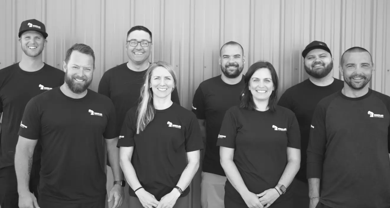 A group of eight people, five men and three women, are standing in a row, smiling at the camera. They are wearing matching black shirts with a small logo on the chest, against a metal corrugated background. The image is in black and white.