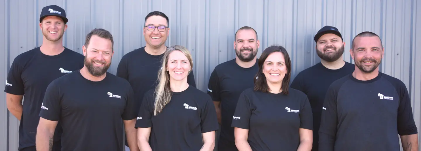 A group of eight people standing in front of a corrugated metal wall. They are wearing matching black shirts with a logo on the chest. There are six men and two women, all smiling.