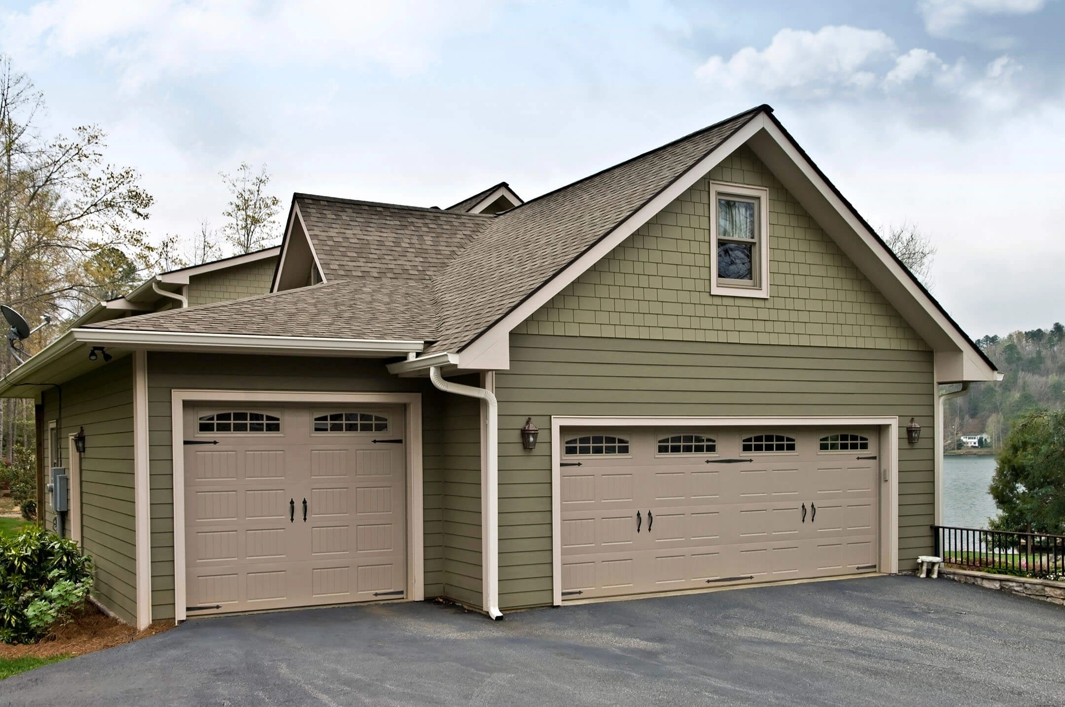 A green house with two beige garage doors and a driveway. The house has a shingled roof and a window above the garages. There are trees and a lake visible in the background under a cloudy sky.