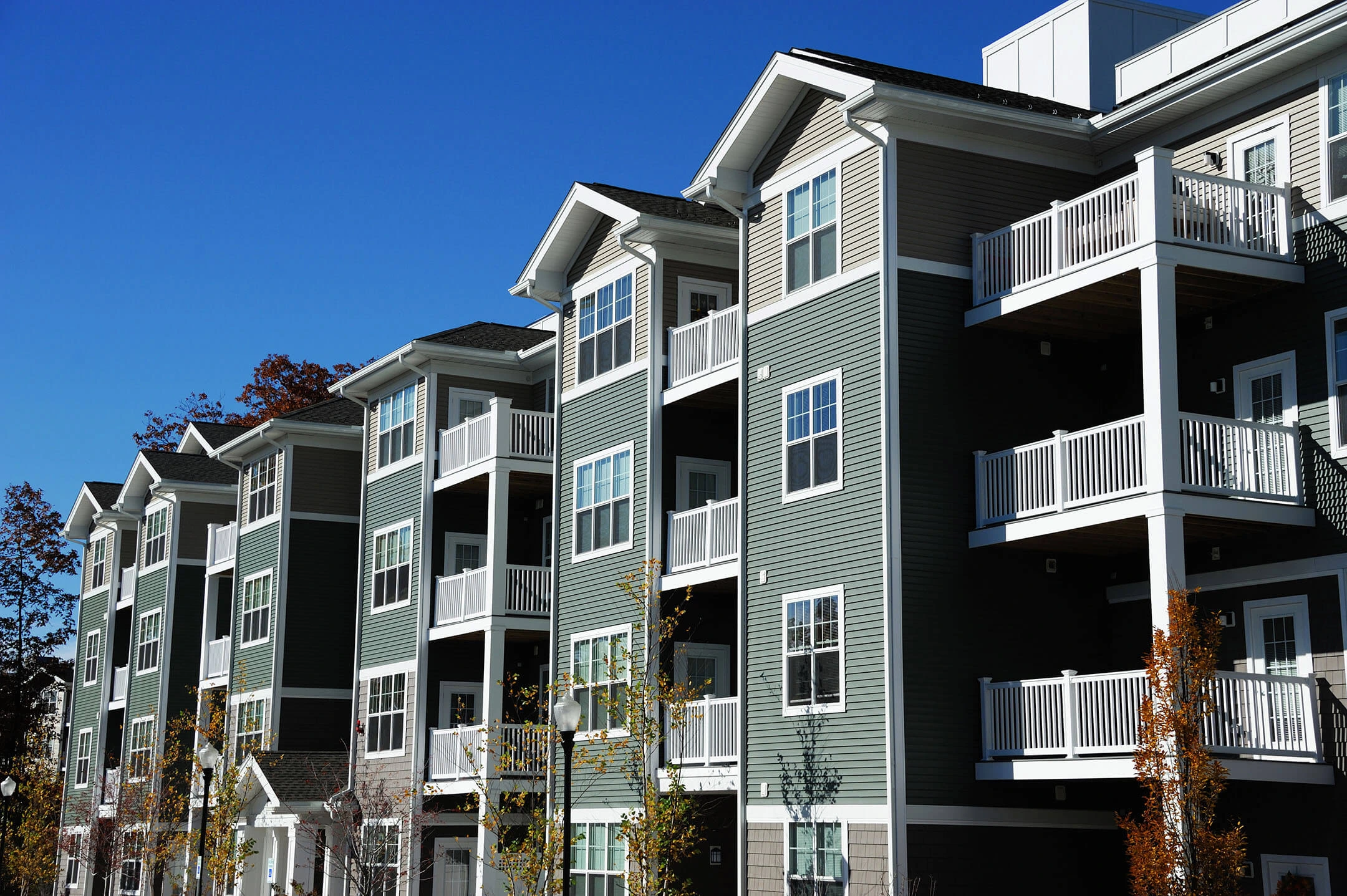 A modern apartment building with green siding and white trim, featuring multiple balconies and large windows. The sky is clear and blue, with a few trees visible in the foreground.