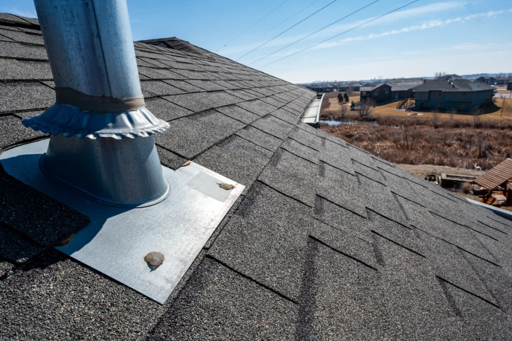 A rooftop with gray asphalt shingles and a metal chimney pipe stands against a clear blue sky, illustrating potential common roofing problems. In the background, a suburban neighborhood with houses and grassy fields is visible.