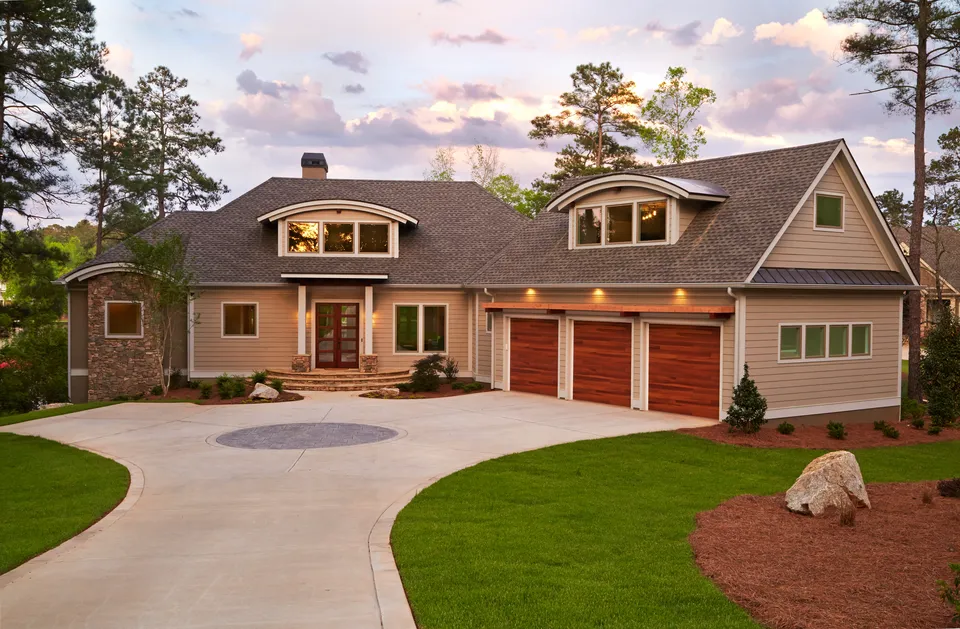 A modern suburban house with a large driveway and three-car garage. The house features stone and wood accents, two dormer windows, and is surrounded by trees and a landscaped garden under a partly cloudy sky.