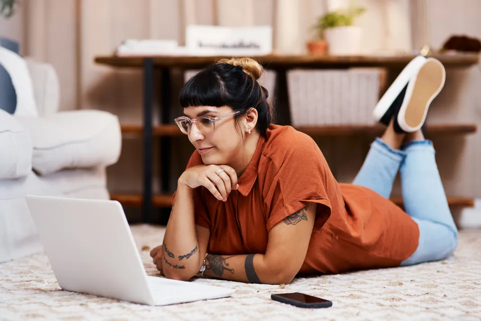 A person with glasses and tattoos, wearing an orange shirt and jeans, is lying on a carpeted floor, propped on their elbows, using a laptop. A smartphone is nearby, and a shelf with decor is in the background.