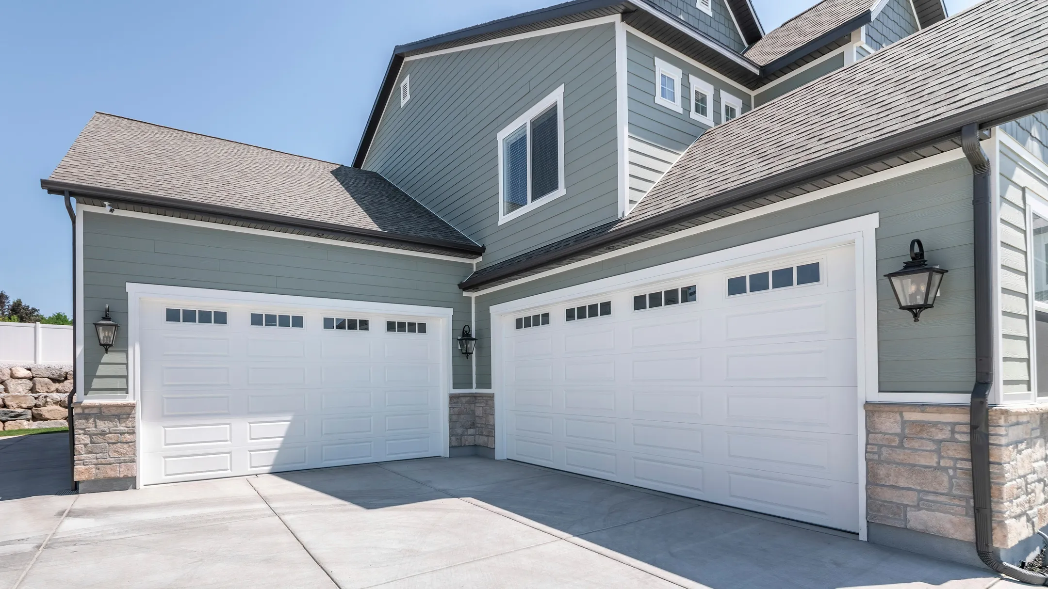 A modern house with light gray and stone exterior features two white garage doors. Each door has four small decorative windows at the top. The driveway is made of smooth concrete, and the house has gray siding with white trim.