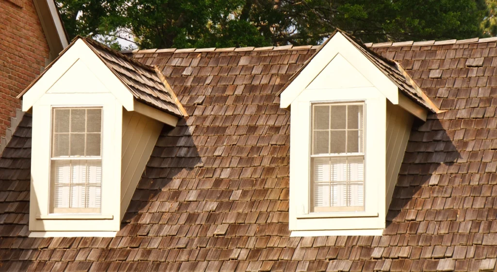 Two dormer windows with white frames and divided panes protrude from a wooden shingle roof, a charming example of various types of roofing. The roof has a warm, sunlit appearance, and green foliage is visible in the background.