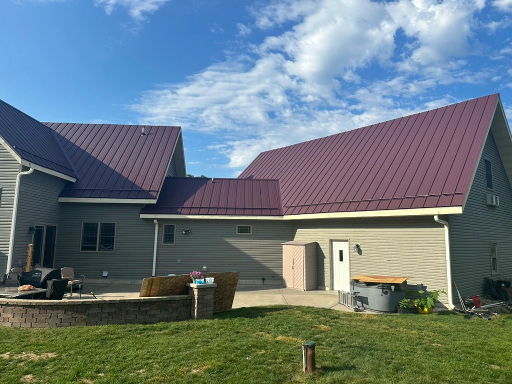 A house with gray siding and a dark red metal roof, set against a blue sky with scattered clouds. There is a patio area with outdoor furniture, a small shed, and a stone fire pit on a green lawn.
