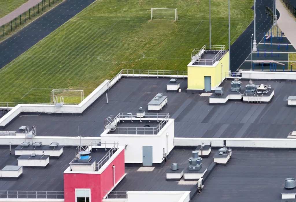 Aerial view of a building rooftop with various structures and equipment. The roof features sections painted in red and yellow. Adjacent to the building, there is a green sports field with a soccer goal and a running track.