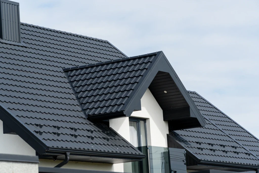 Close-up of a modern house roof with black, metal tiles and a triangular dormer. The sleek design not only enhances its contemporary architectural style against a cloudy sky but also offers an insight into potential metal roof cost considerations.