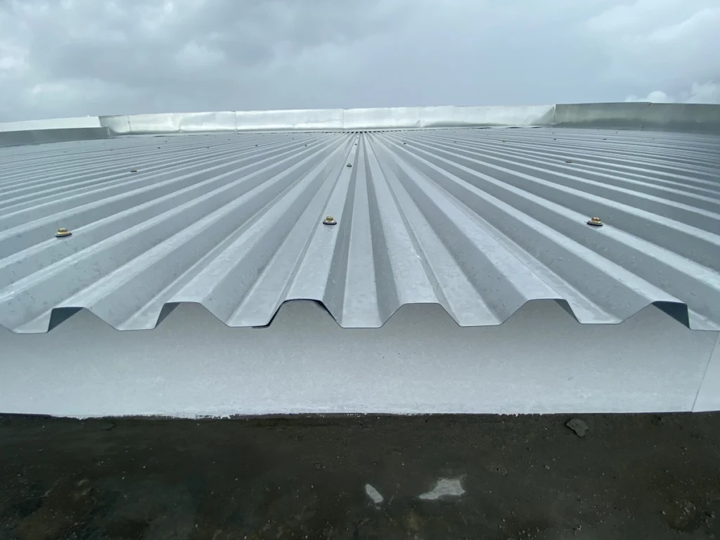 A view of corrugated metal roofing stretches beneath a cloudy sky, its ridges and grooves forming a linear pattern. Bolts visibly secure the sheets, while the ground peeks through at the bottom, adding depth to this industrial scene.