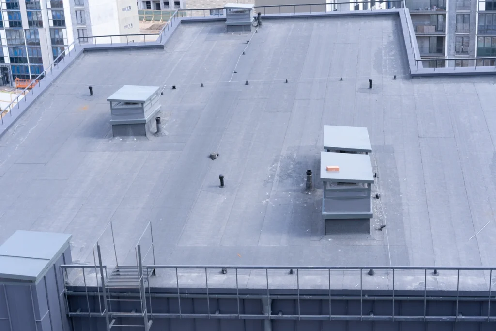 Flat rooftop of a city building with two rectangular vent structures. The surface is gray and features scattered pipes and a book on one vent. Surrounding the roof are taller buildings with visible windows and balconies.