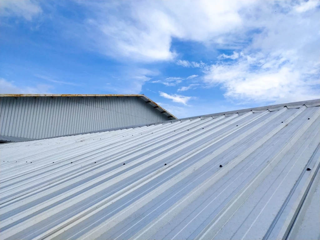 A close-up view of corrugated metal roofing under a blue sky with scattered clouds. The roof's horizontal lines lead toward the horizon, creating a sense of depth. The bright sky contrasts with the silvery, reflective surface of the corrugated material.
