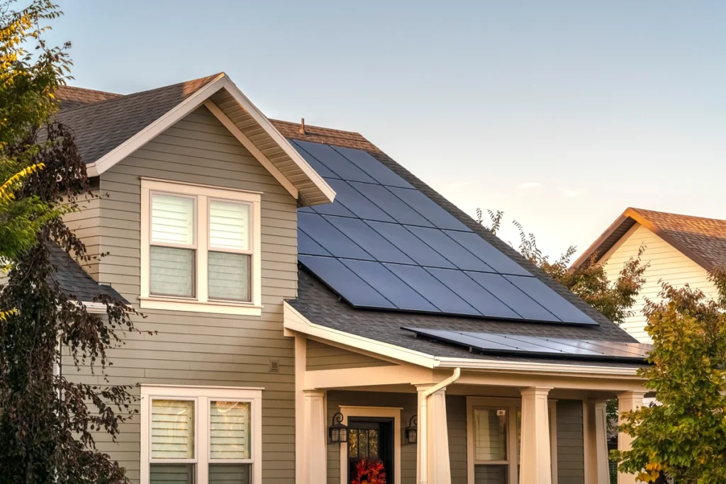 A modern two-story house with gray siding boasts solar panels on the roof, hinting at sustainability. Surrounded by trees and bathed in late afternoon sunlight, it evokes tranquility. The clear sky glows warmly as you ponder, how long do solar panels last while enjoying the serene view?.